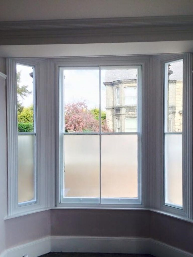 Close-up of a white-framed secondary glazed window installed inside an older wooden window frame, showing double glazing panels and latch mechanism, with natural light visible through the glass.